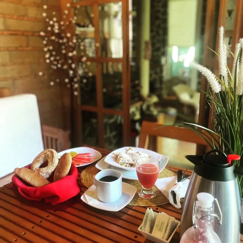 Mesa de desayuno completa: canasta de pan dulce, fruta, café de olla, licuado y jarra de agua fresca junto a la ventana