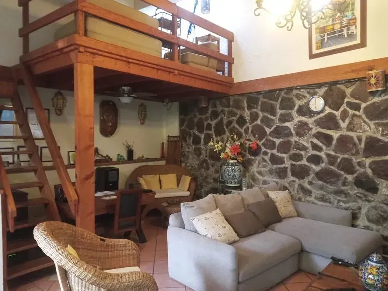 Sala común con muro de piedra volcánica, sofá de esquina, sillón de mimbre y tapanco de madera con escalera