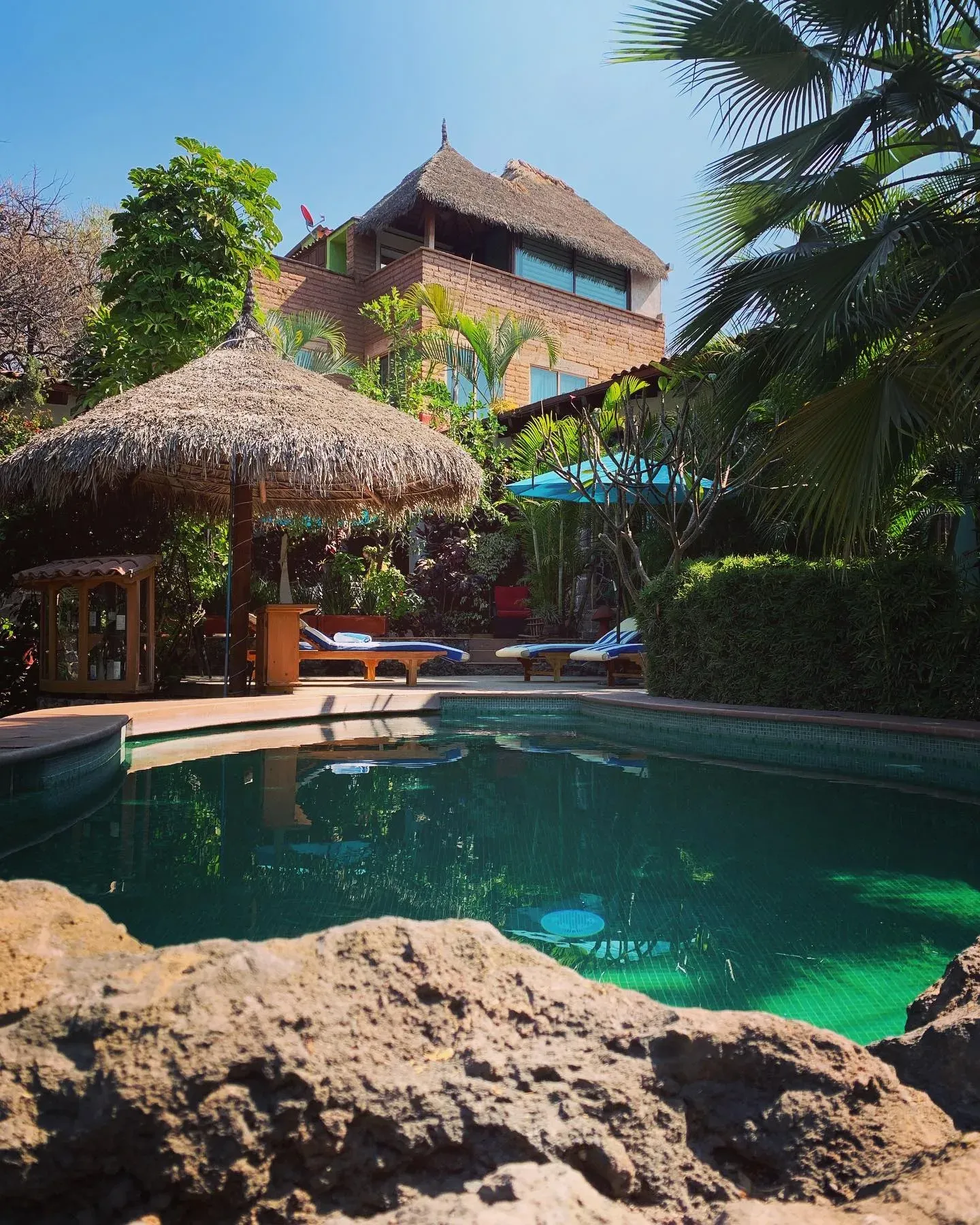 Alberca de agua verde con palapa de palma y camastros, la casa principal de adobe al fondo entre palmeras y cielo azul