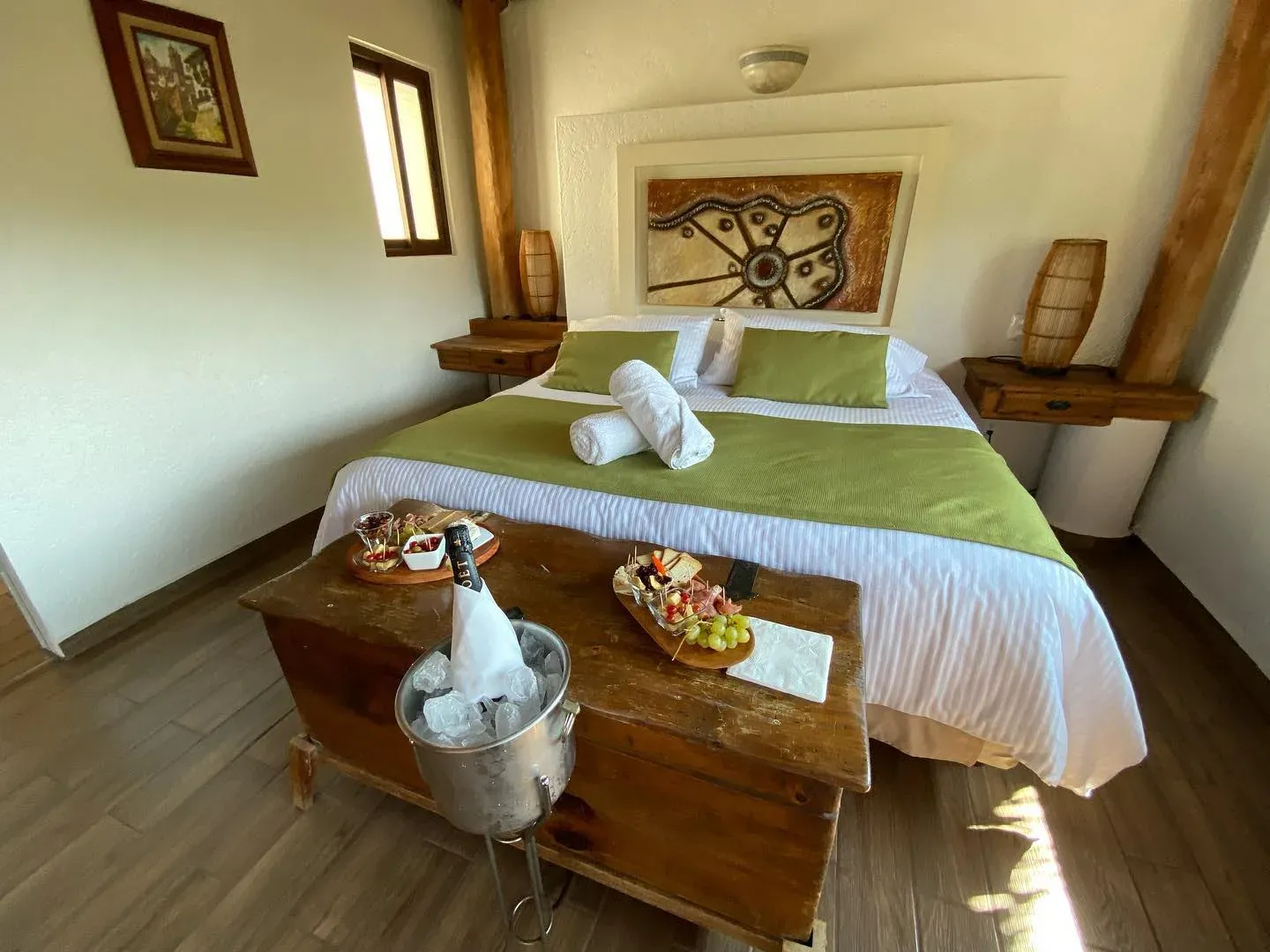 Cama con textiles verdes y toallas dobladas, baúl de madera con fruta y hielera, bajo luz cálida de tarde