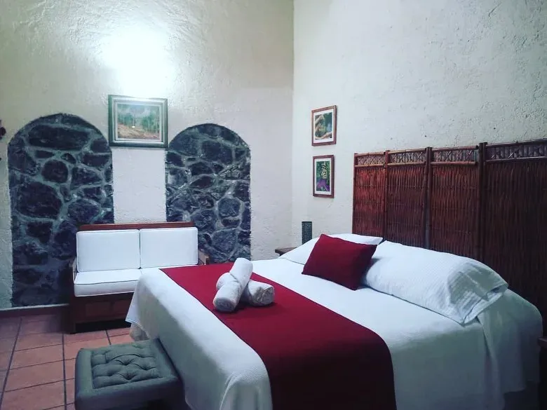 Habitación de techo alto con dos nichos de piedra volcánica, cabecera de madera labrada y camino rojo sobre la cama